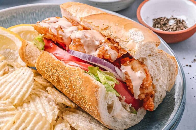 Close-up of a single shrimp po’boy sandwich on a plate with chips. The sandwich is drizzled with creamy sauce and packed with crispy shrimp, fresh tomato, lettuce, and red onion.