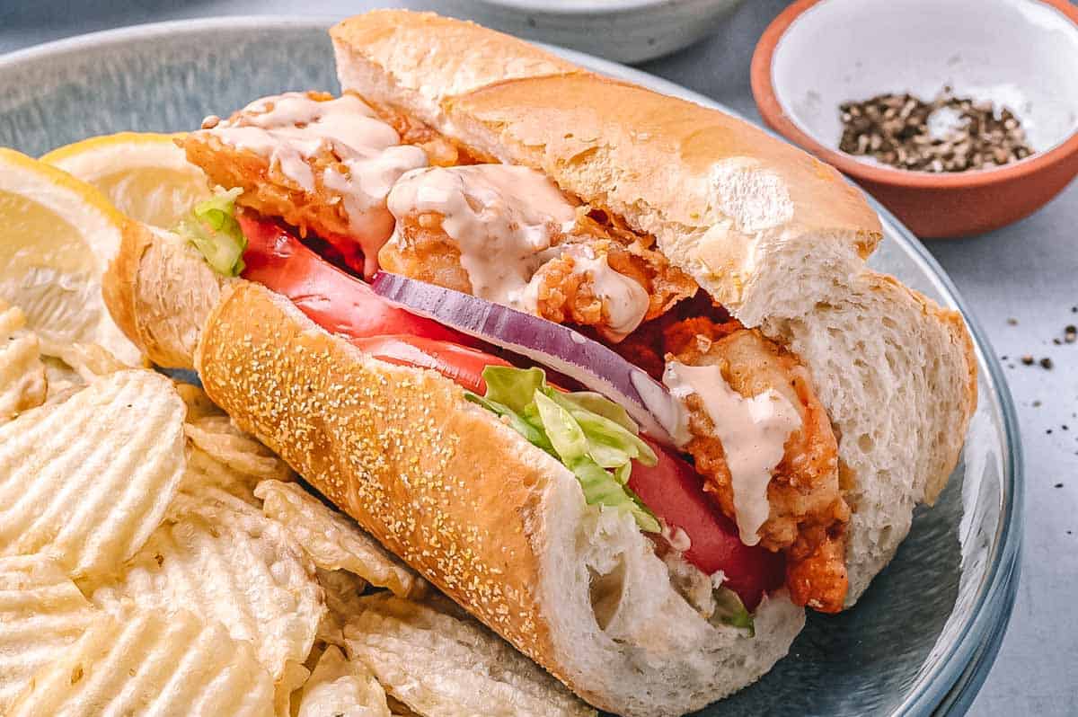 Close-up of a single shrimp po’boy sandwich on a plate with chips. The sandwich is drizzled with creamy sauce and packed with crispy shrimp, fresh tomato, lettuce, and red onion.
