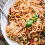 A close-up of spaghetti dressed in a chunky tomato sauce, garnished with a fresh basil leaf. The pasta is lightly tossed with finely chopped vegetables and nuts, and a fork is gently twirled into a portion, ready to be lifted.