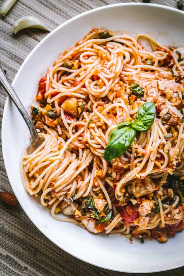 A close-up of spaghetti dressed in a chunky tomato sauce, garnished with a fresh basil leaf. The pasta is lightly tossed with finely chopped vegetables and nuts, and a fork is gently twirled into a portion, ready to be lifted.