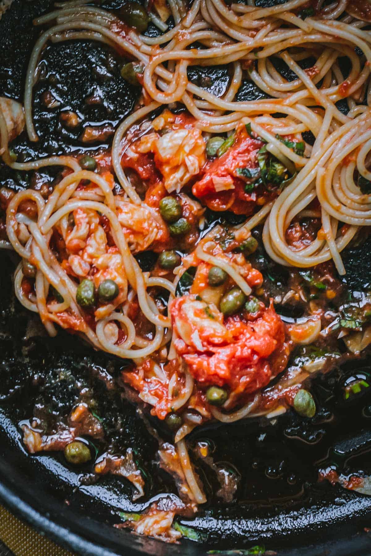 Spaghetti glistens in a deep, rustic skillet, coated in a rich tomato sauce with visible chunks of fish and plump capers. The pan’s dark surface highlights the vibrant reds and greens of the dish’s fresh ingredients.