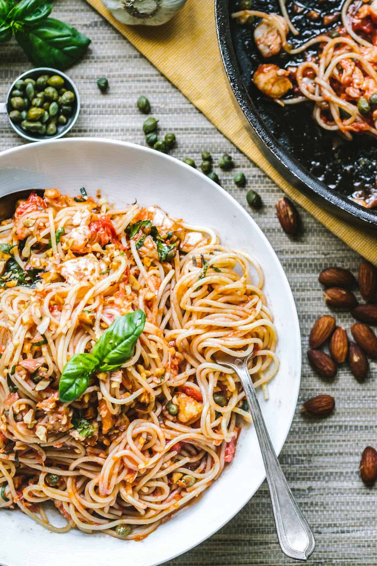 A white bowl brimming with saucy spaghetti sits at the center of a cozy tabletop. Surrounding it are raw almonds, capers, and garlic cloves—hinting at the Mediterranean ingredients used in the dish.