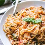 A warm-toned photo of spaghetti twirled on a fork, resting in a bowl of tomato-coated noodles. Chopped basil and pieces of fish are nestled among the strands, delivering a sense of freshness and texture.