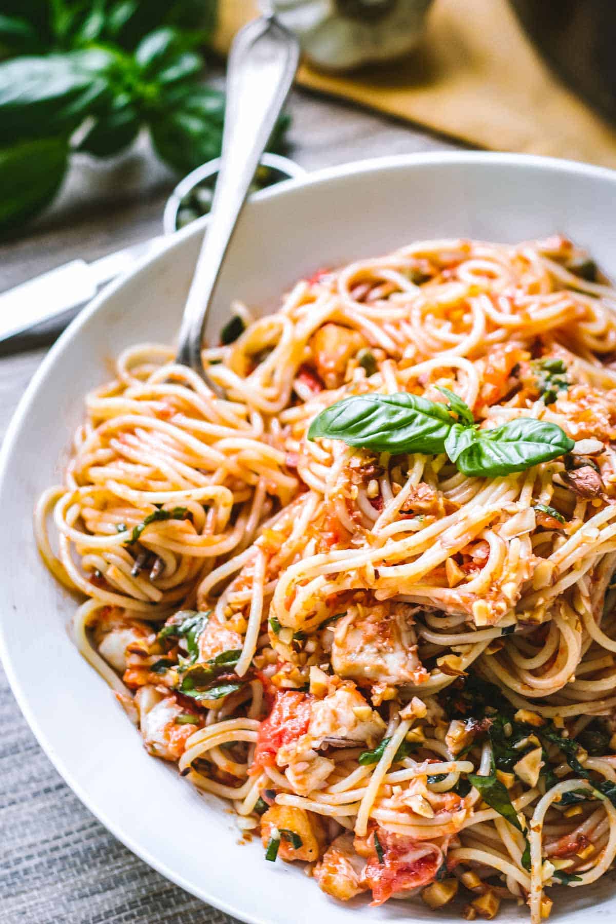 A warm-toned photo of spaghetti twirled on a fork, resting in a bowl of tomato-coated noodles. Chopped basil and pieces of fish are nestled among the strands, delivering a sense of freshness and texture.