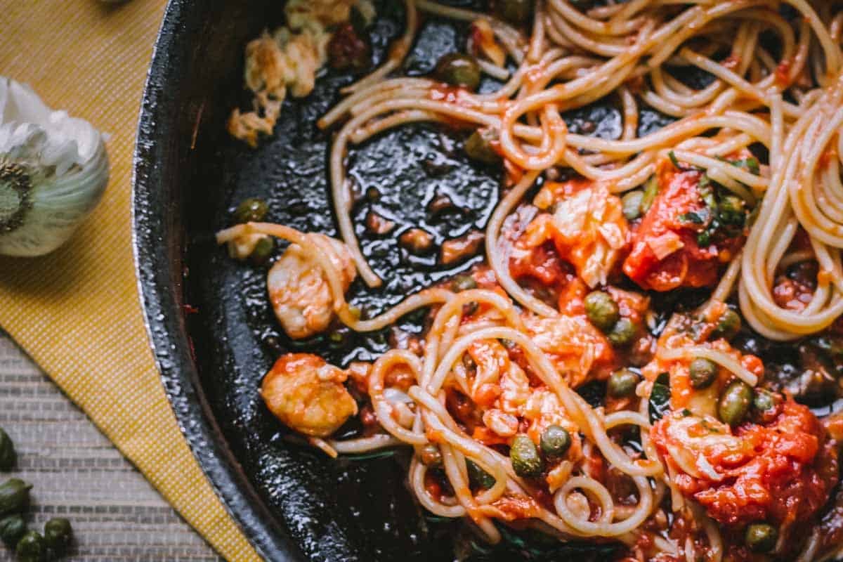 A rustic skillet holds the last saucy bites of spaghetti, with visible capers and tomato chunks still glistening. The remnants offer a glimpse into a homey, satisfying meal made with bold, briny flavors.