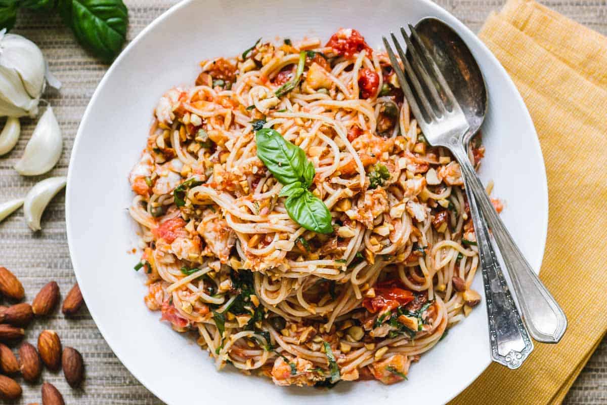 A composed flat lay of a pasta dish topped with basil, served in a clean white bowl alongside vintage silverware. The scene is accented by whole almonds, peeled garlic cloves, and a mustard-colored napkin, evoking a home-cooked feel.