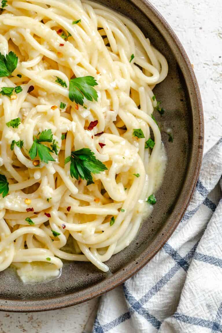 Close-up detail of pasta aglio e olio in a ceramic bowl, highlighting how the simple sauce clings to each strand of spaghetti. The parsley and red pepper flakes provide visual contrast against the pasta, and a fork twirling through the noodles demonstrates the dish's perfect consistency.