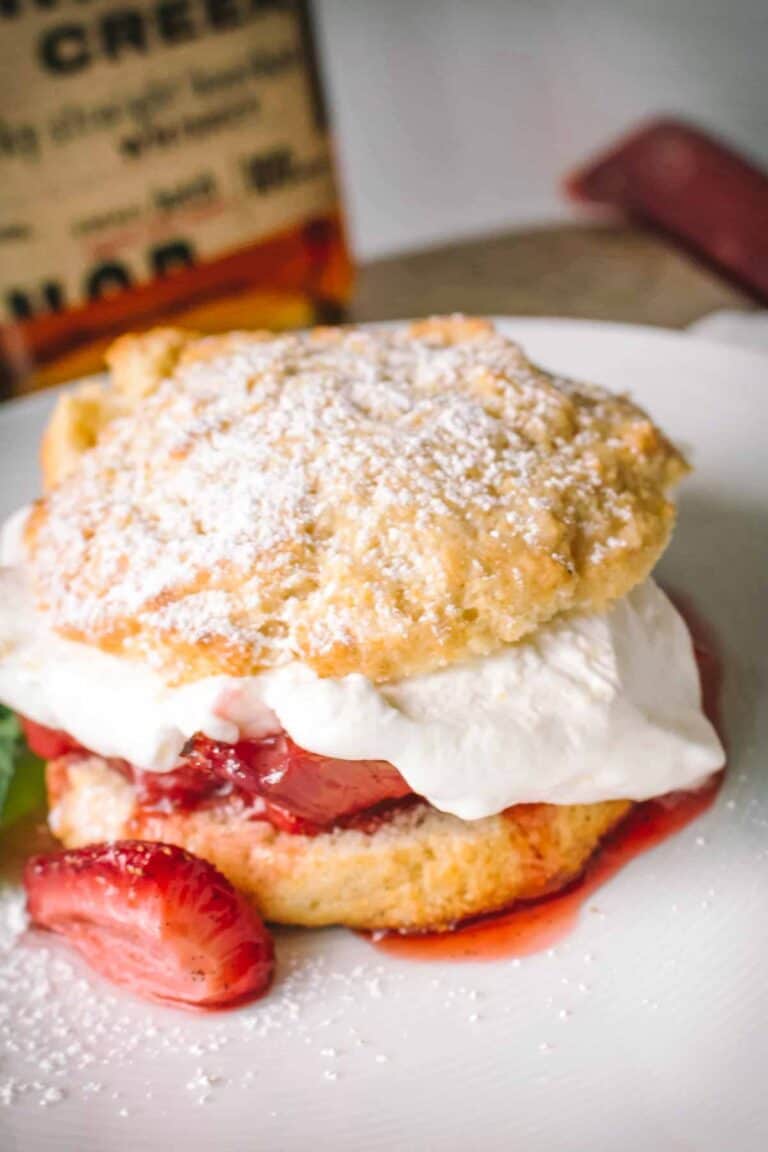 Close-up of a split shortcake biscuit topped with whipped cream and fresh strawberries, dusted with powdered sugar. A bottle of whiskey is partially visible in the background, suggesting a boozy addition to the dessert.