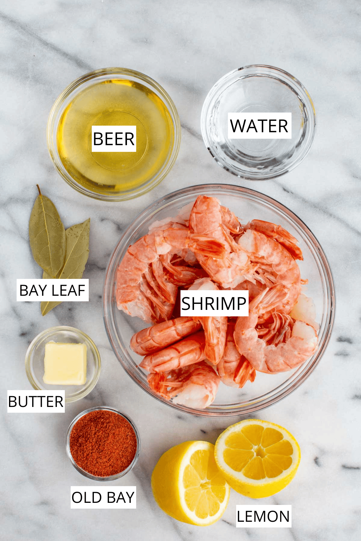 Flat lay of the ingredients for peel-and-eat shrimp: raw shrimp, halved lemons, bay leaves, butter, beer, water, Old Bay, and olive oil arranged on a marble surface.