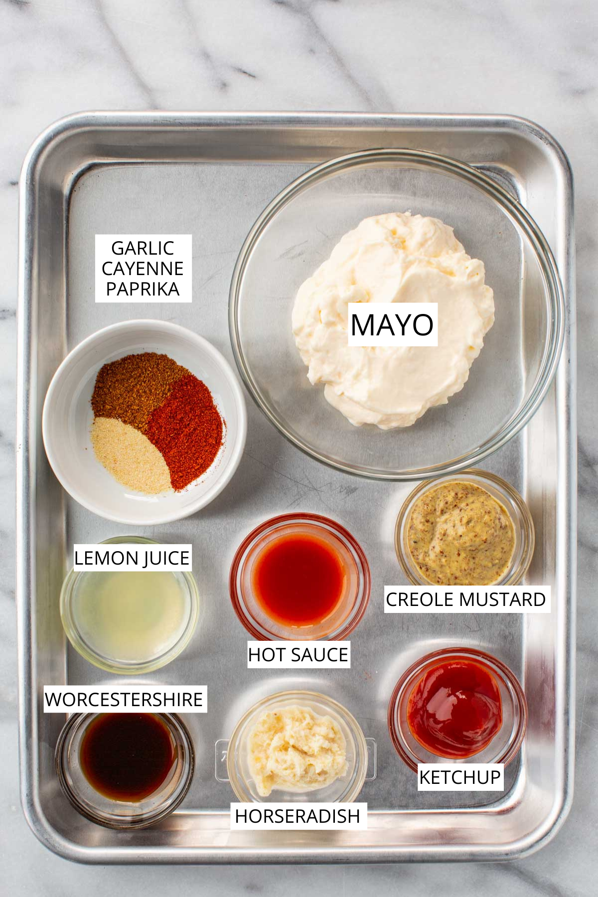 Labeled ingredients for Creole remoulade sauce displayed on a metal tray, including a large bowl of mayonnaise, a bowl of ketchup, prepared horseradish, Creole mustard, hot sauce, lemon juice, Worcestershire sauce, and a small dish with garlic powder, cayenne, and paprika.
