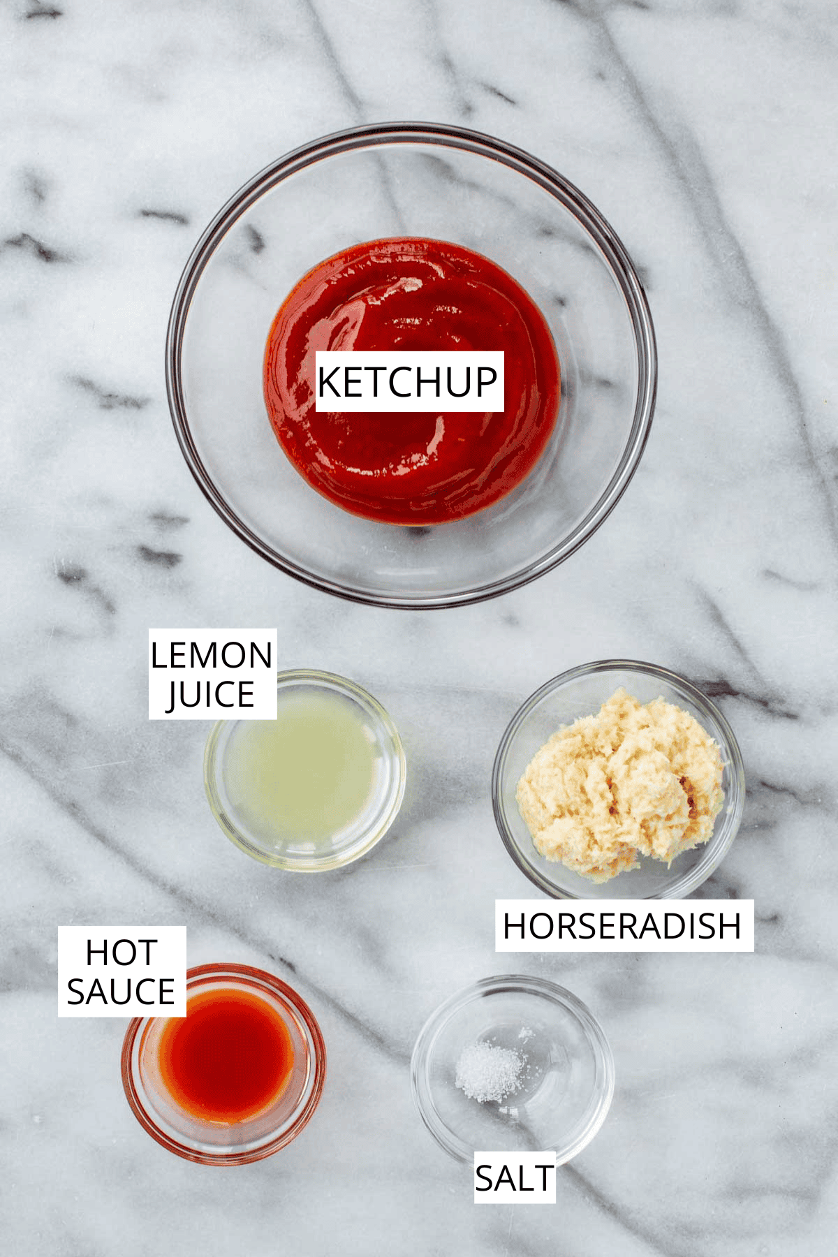 Labeled ingredients for cocktail sauce displayed on a marble surface: ketchup in a glass bowl, lemon juice, hot sauce, prepared horseradish, and salt in small glass containers.