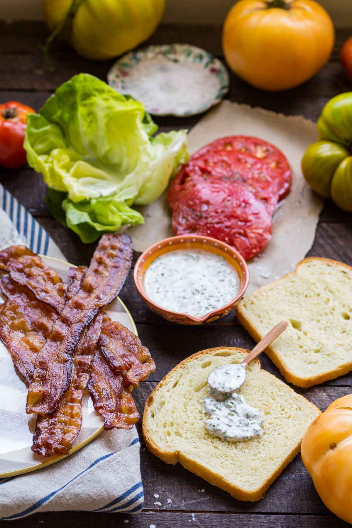 Crispy bacon, lettuce, tomato slices, herb mayo, and white bread laid out for assembling a BLT sandwich.