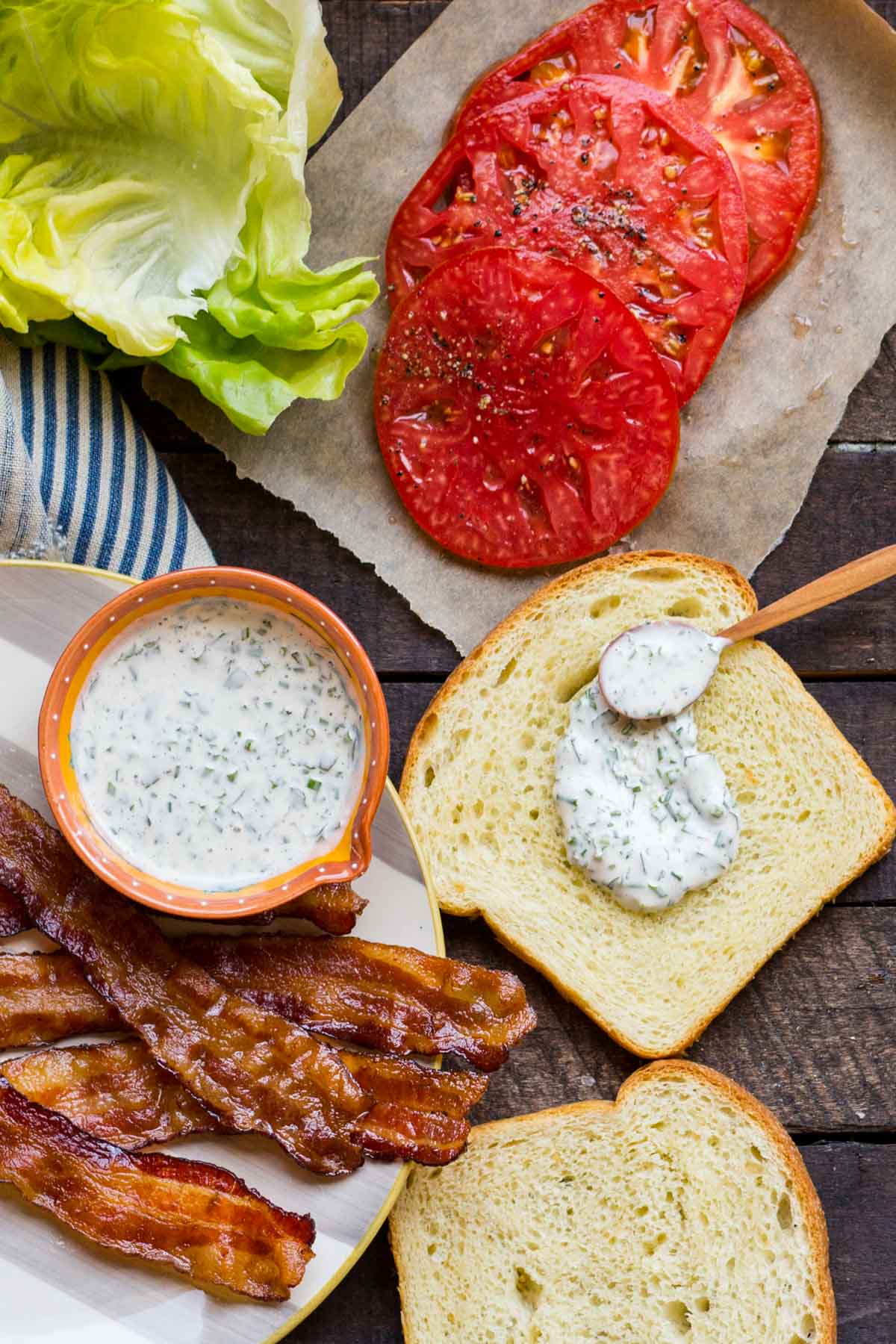 Lettuce, tomatoes, bacon, mayo, and bread arranged on a dark wood surface, with mayo spread on one slice.