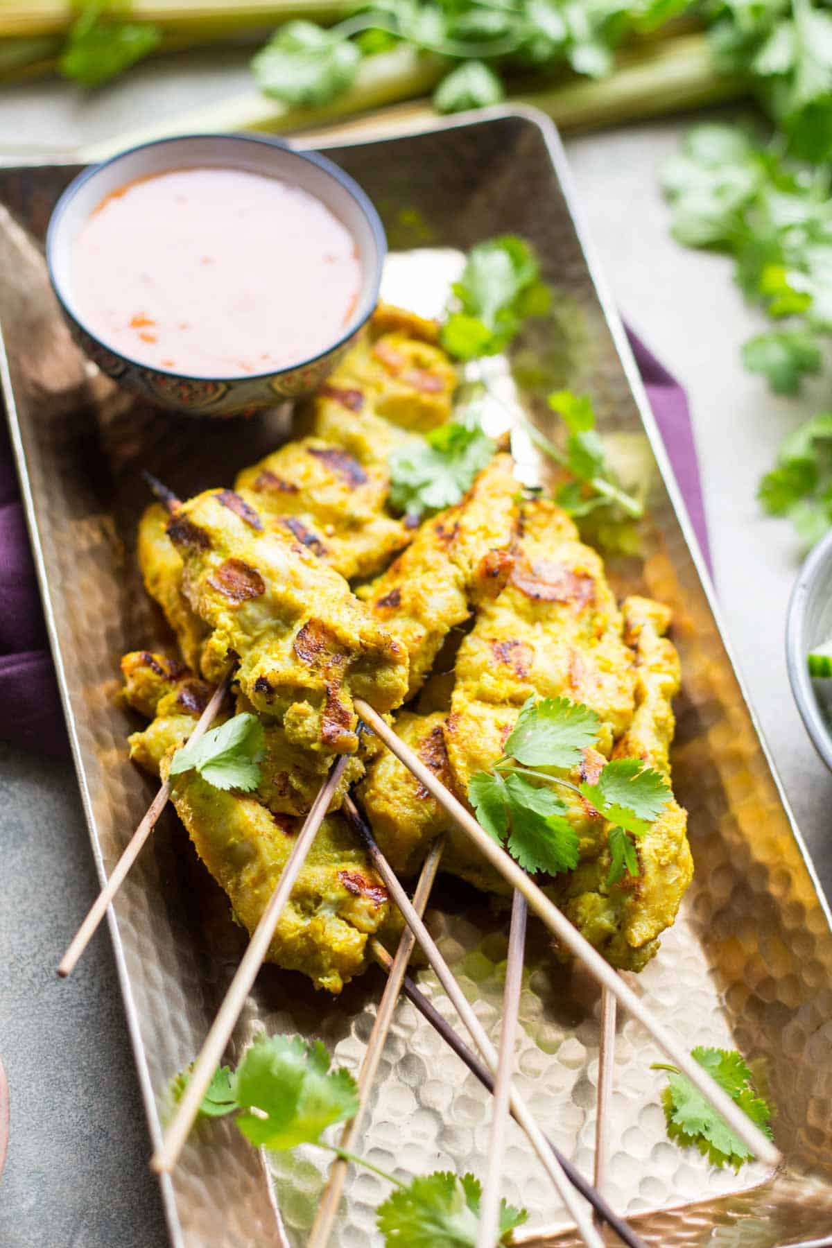 Grilled chicken satay skewers arranged on a hammered copper tray, garnished with fresh cilantro and served with a small bowl of creamy peanut dipping sauce.