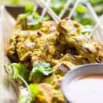 Close-up view of char-grilled chicken satay skewers, with golden turmeric marinade and cilantro, with a bowl of peanut sauce partially visible in the foreground.