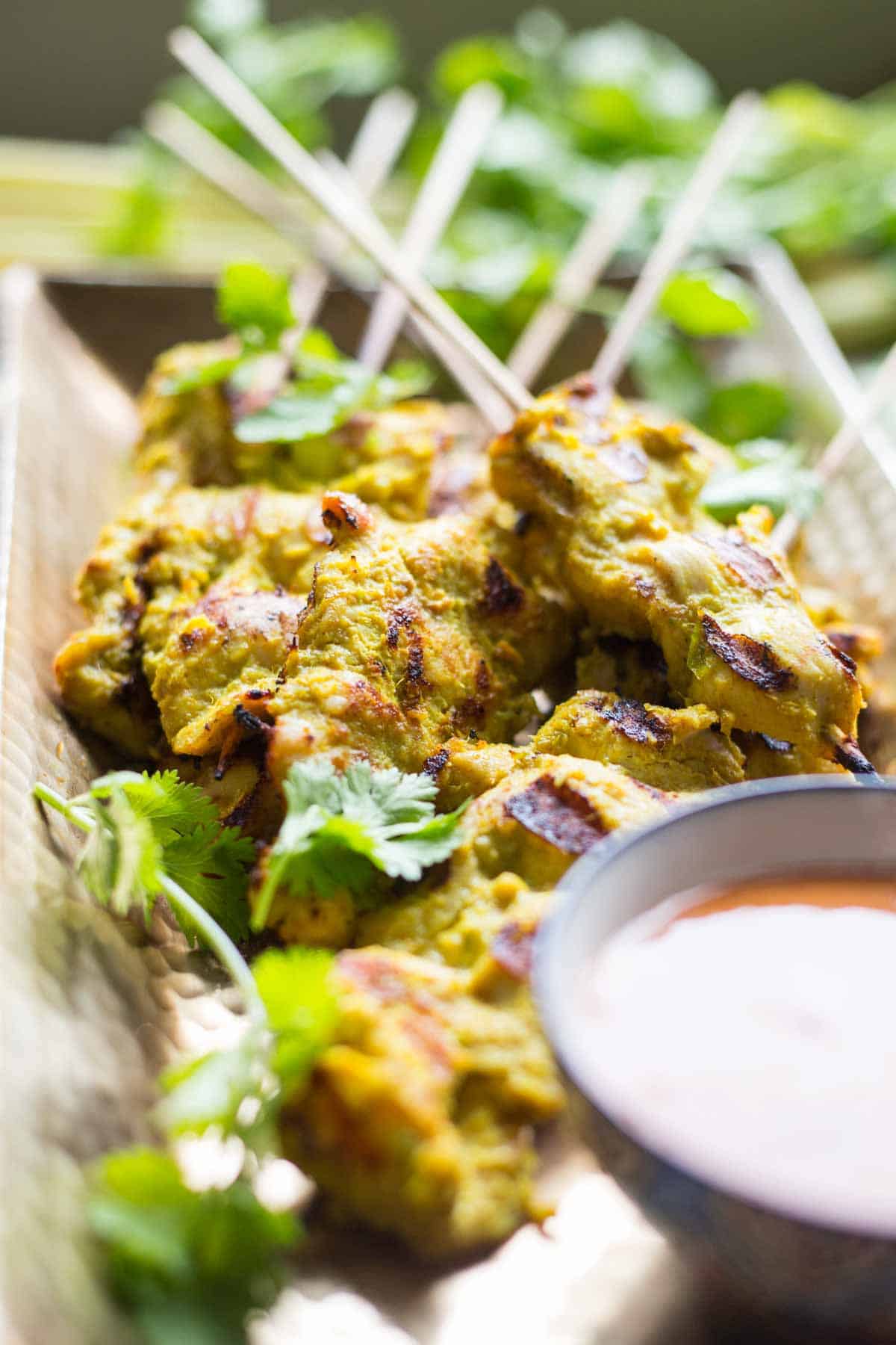 Close-up view of char-grilled chicken satay skewers, with golden turmeric marinade and cilantro, with a bowl of peanut sauce partially visible in the foreground.