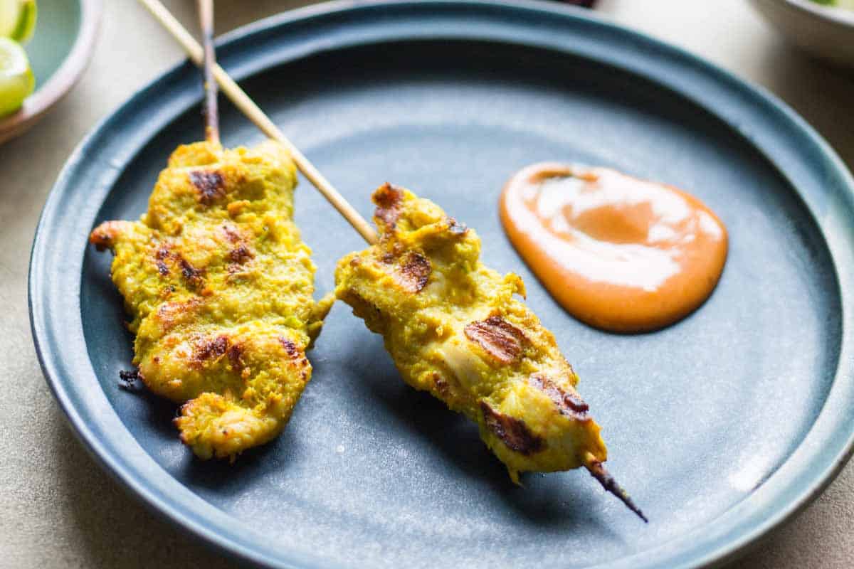 Two chicken satay skewers on a matte blue plate served with a dollop of peanut sauce, highlighting the seared grill marks on the meat.