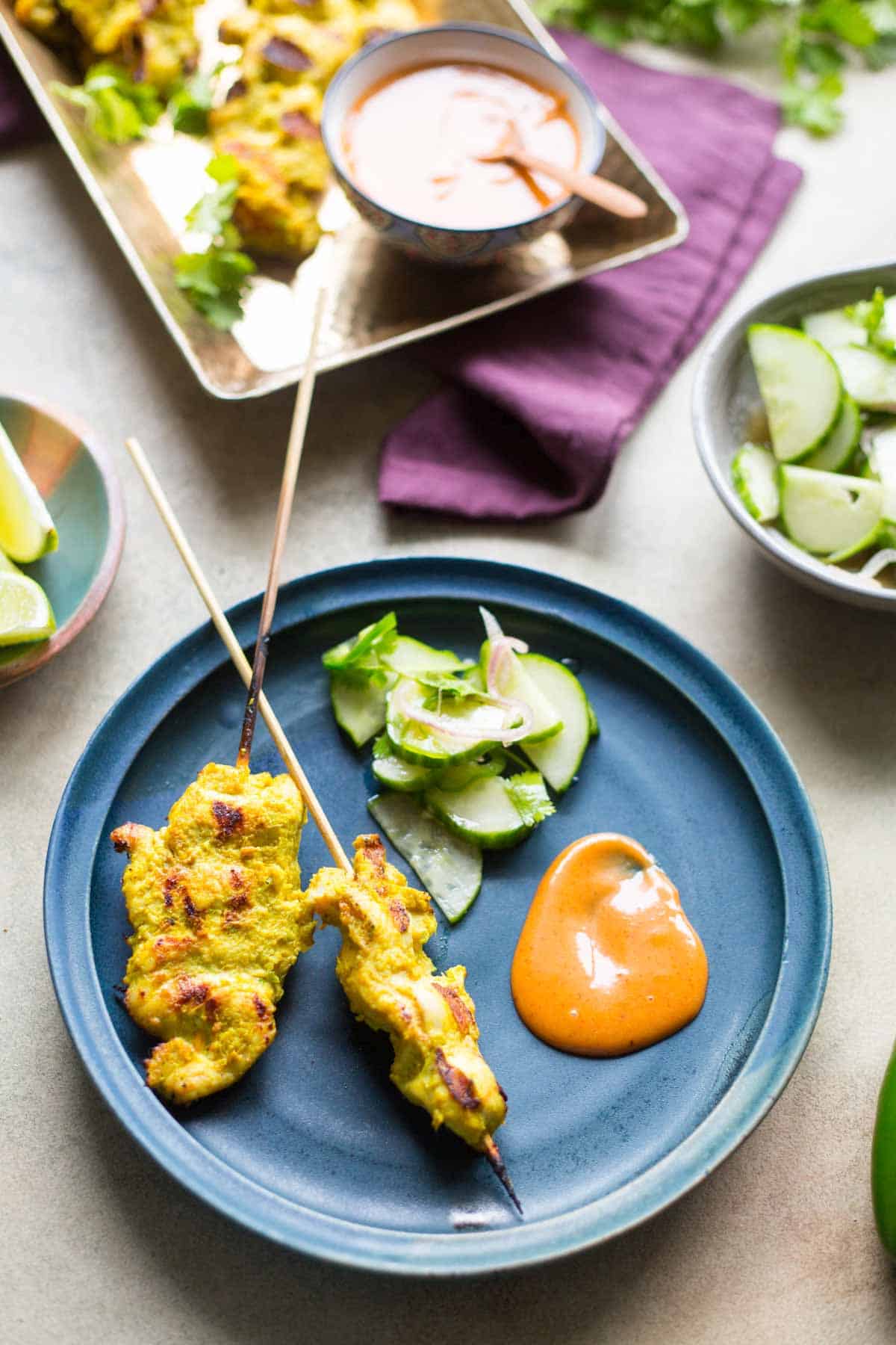 A top-down plate view featuring two chicken satay skewers, a mound of creamy peanut sauce, and a side of quick cucumber salad with herbs and shallots.