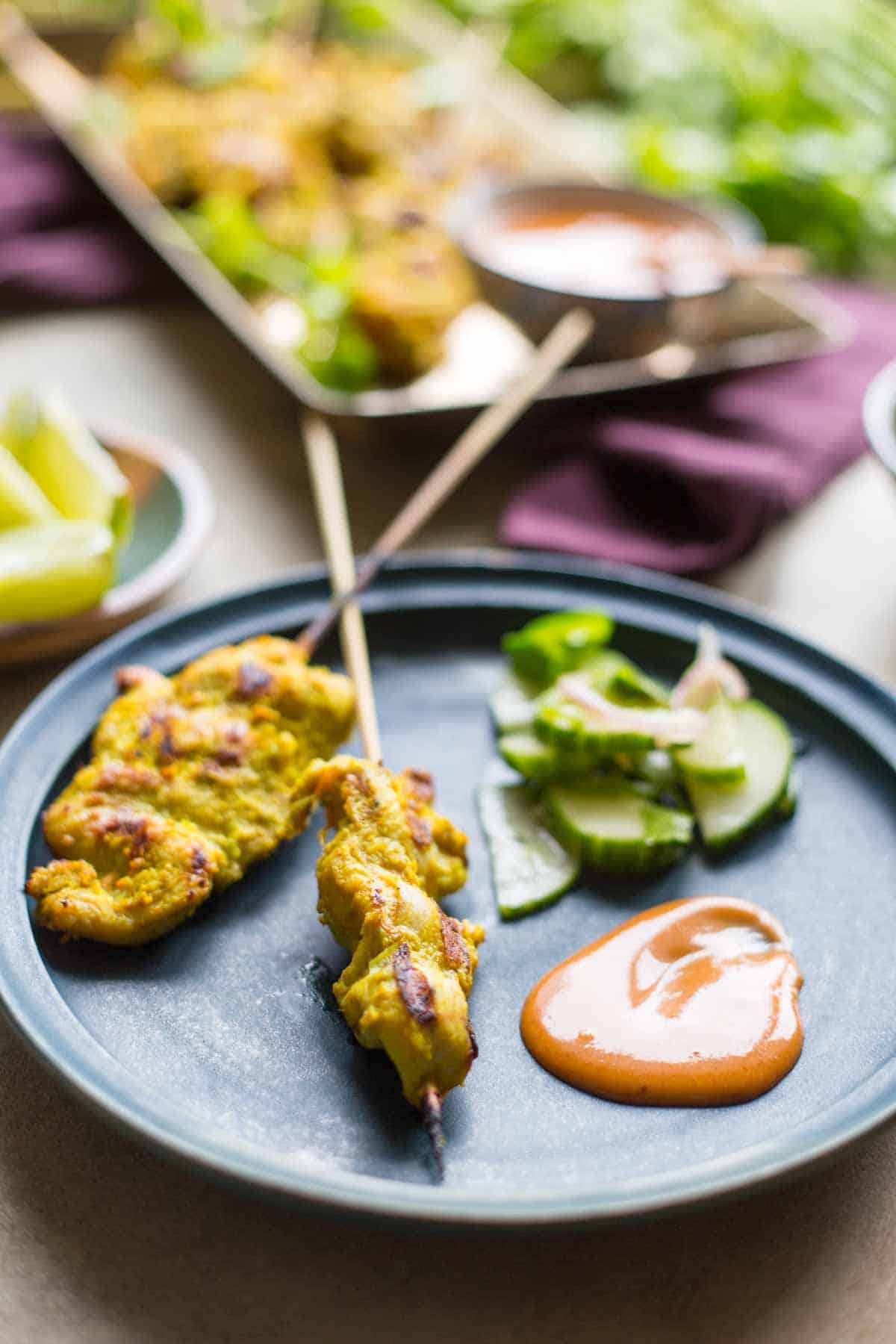 Side angle of plated chicken satay skewers with a fresh cucumber salad and peanut sauce, with the serving tray and dipping bowl softly blurred in the background.
