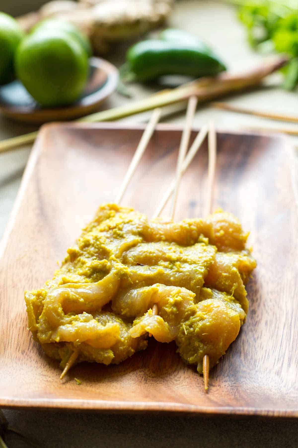 Raw chicken pieces coated in golden lemongrass marinade threaded onto bamboo skewers and arranged on a wooden tray, with jalapeños and fresh herbs in the background.