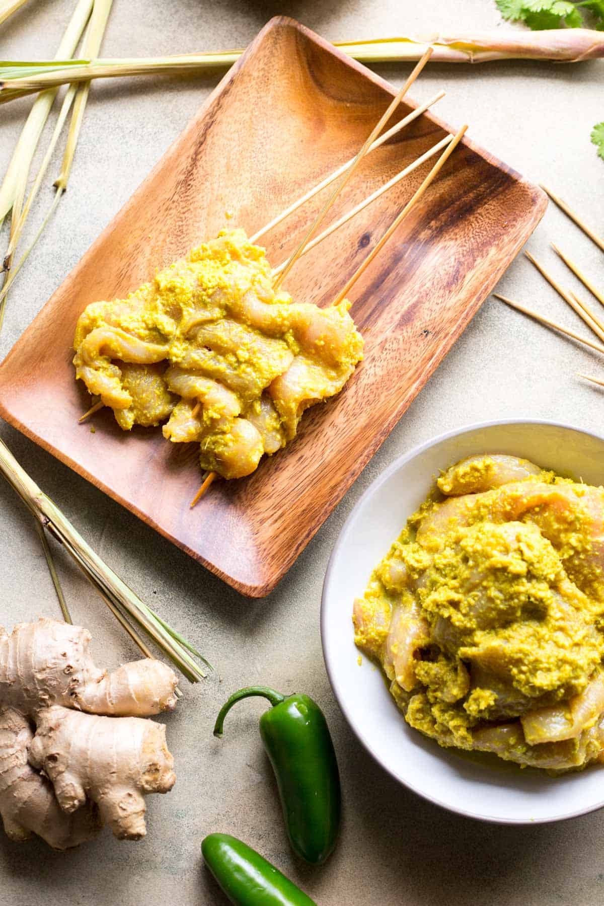 Overhead shot of marinated raw chicken skewers on a wood tray next to a white bowl of additional chicken in marinade, surrounded by lemongrass, ginger, and green chili peppers.