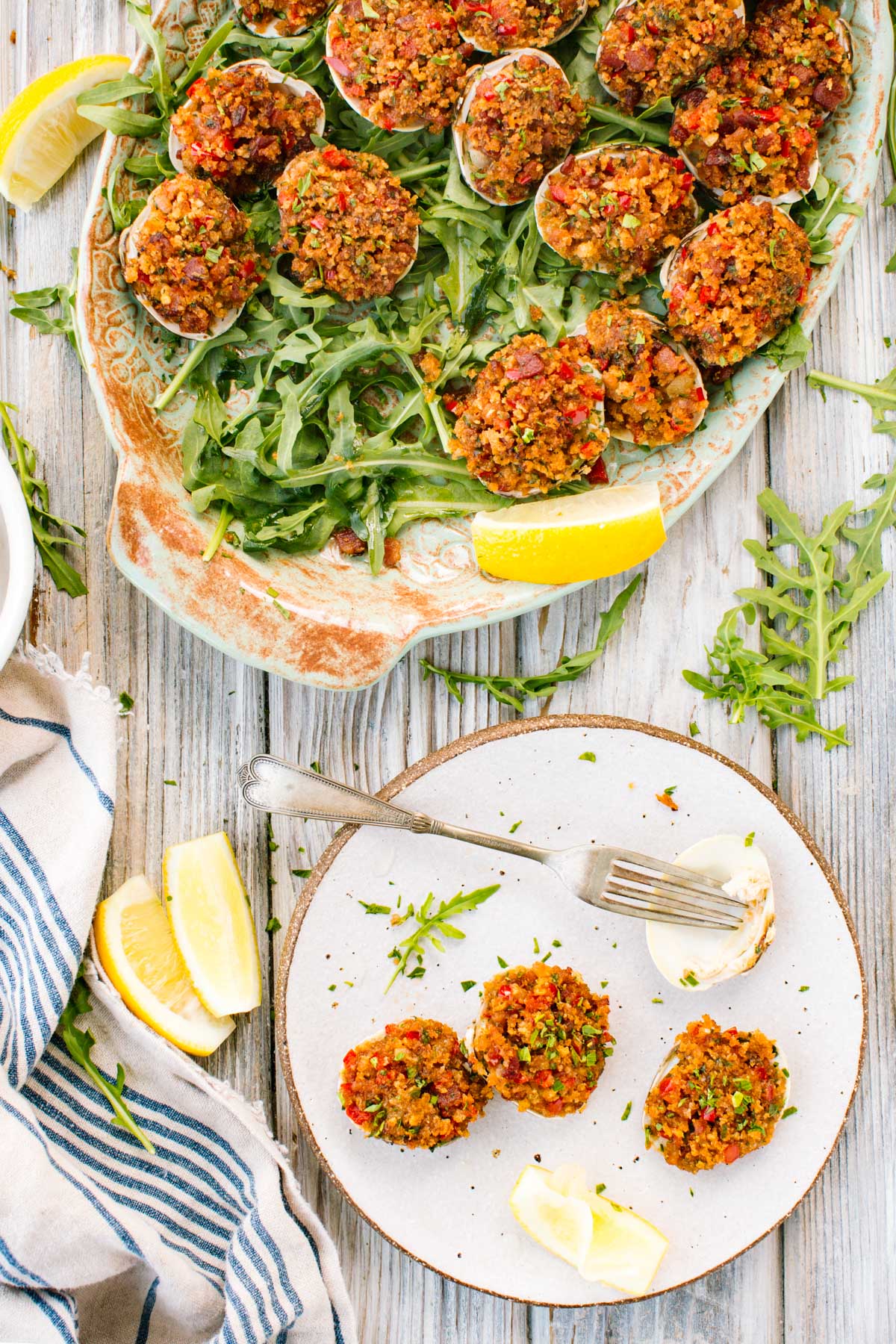 Stuffed clams on a platter with greens and lemon wedges, plus a small plate with three clams and a fork.