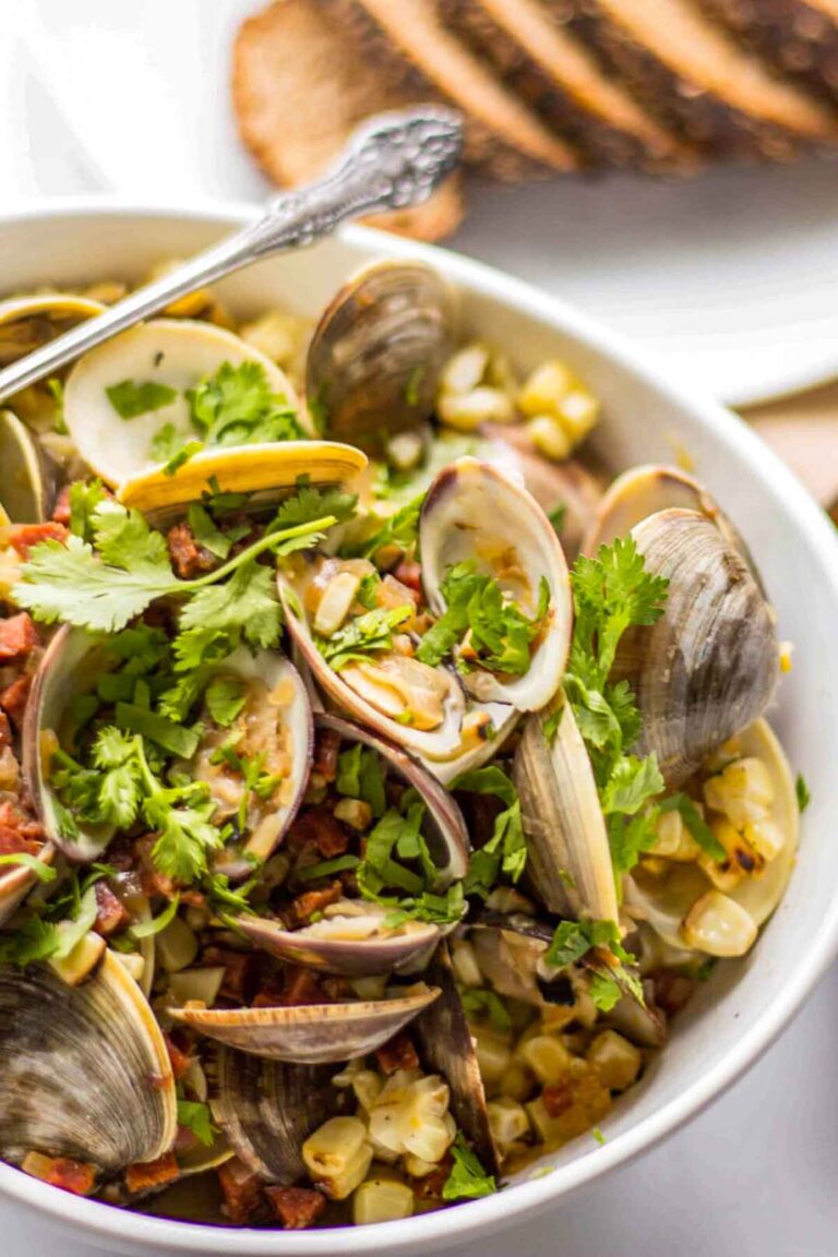 Clams in a white bowl with charred corn, chorizo, and herbs, served with rustic bread.