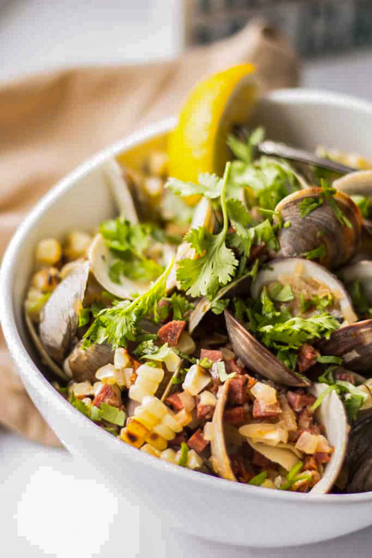 Clams tossed with chorizo, corn, and cilantro, garnished with a lemon wedge in a white bowl.