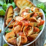 Angled close-up of the main bowl, emphasizing the vibrant tomato sauce, opened clams, fresh herbs, and melted blue cheese, with a silver fork resting in the bowl.