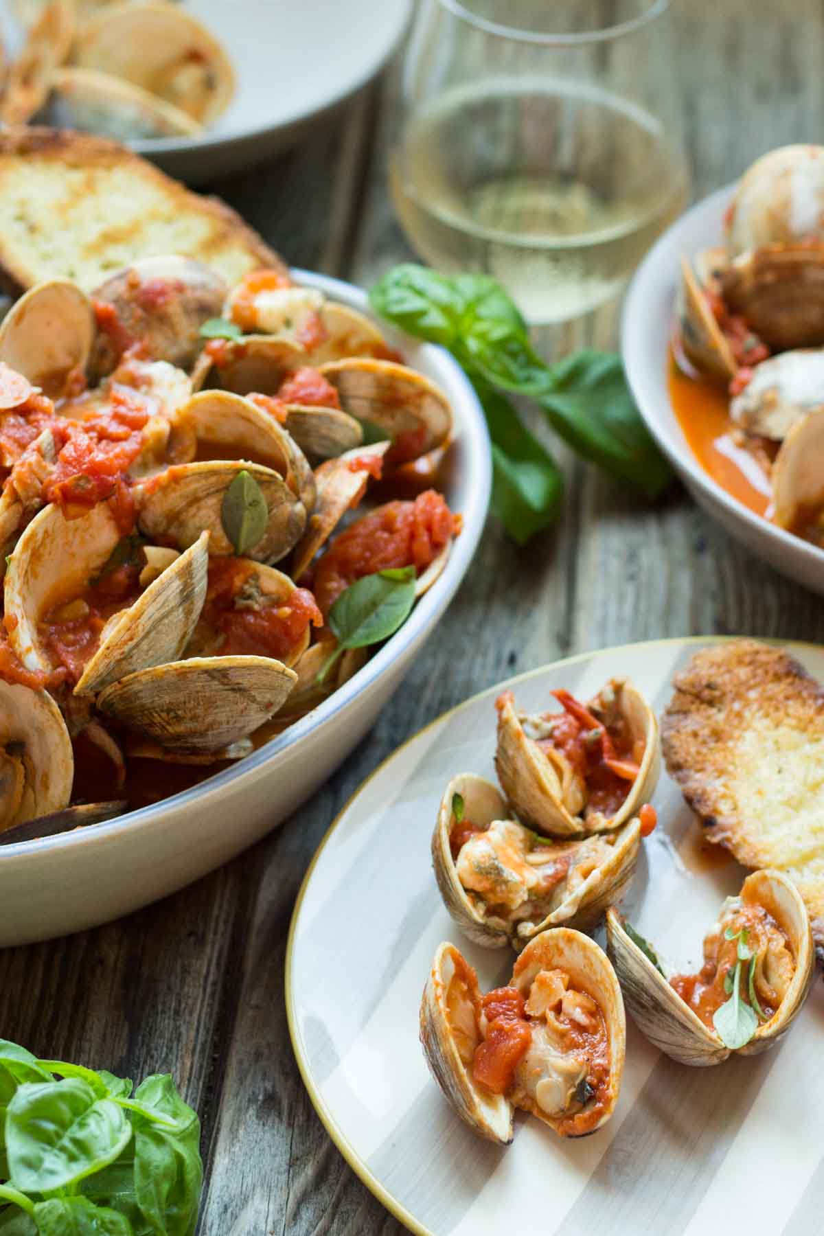 Close-up of clams on a plate topped with tomato sauce and herbs, next to a crispy piece of grilled bread.