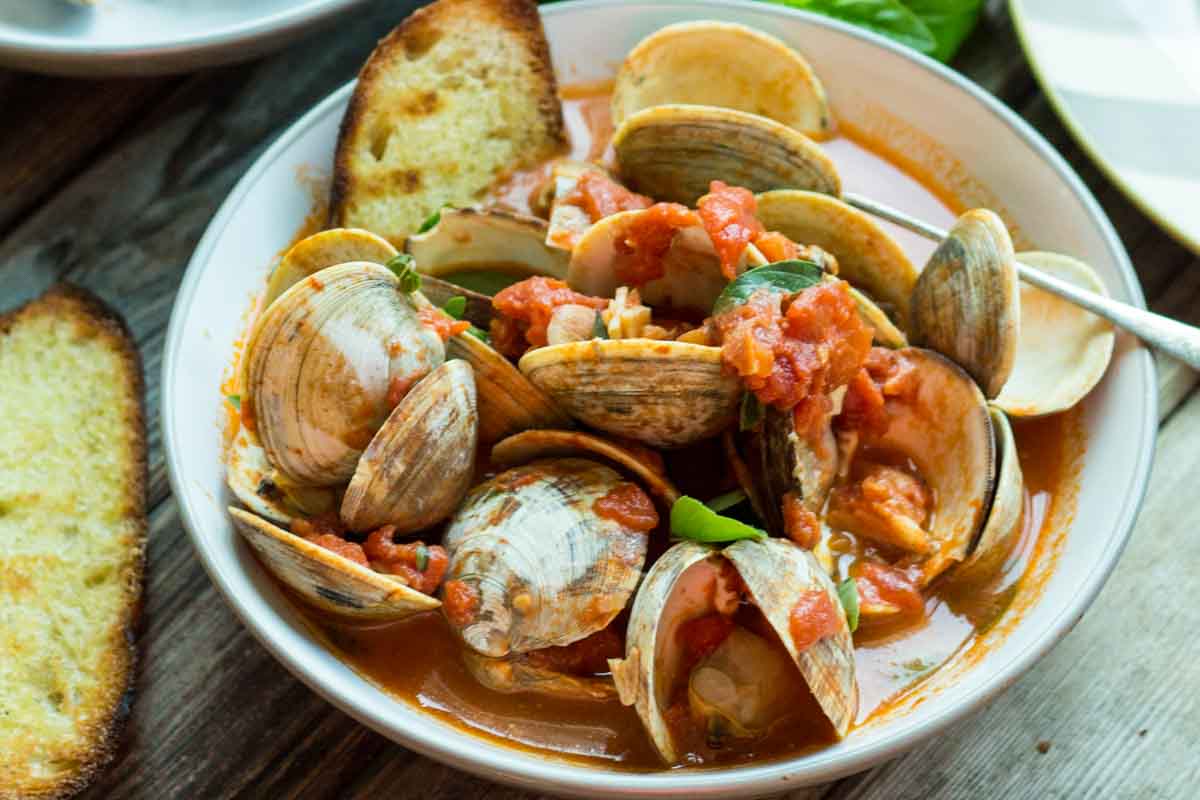 Side view of a bowl of clams in tomato broth, garnished with fresh basil and crumbles of blue cheese, served with grilled bread.