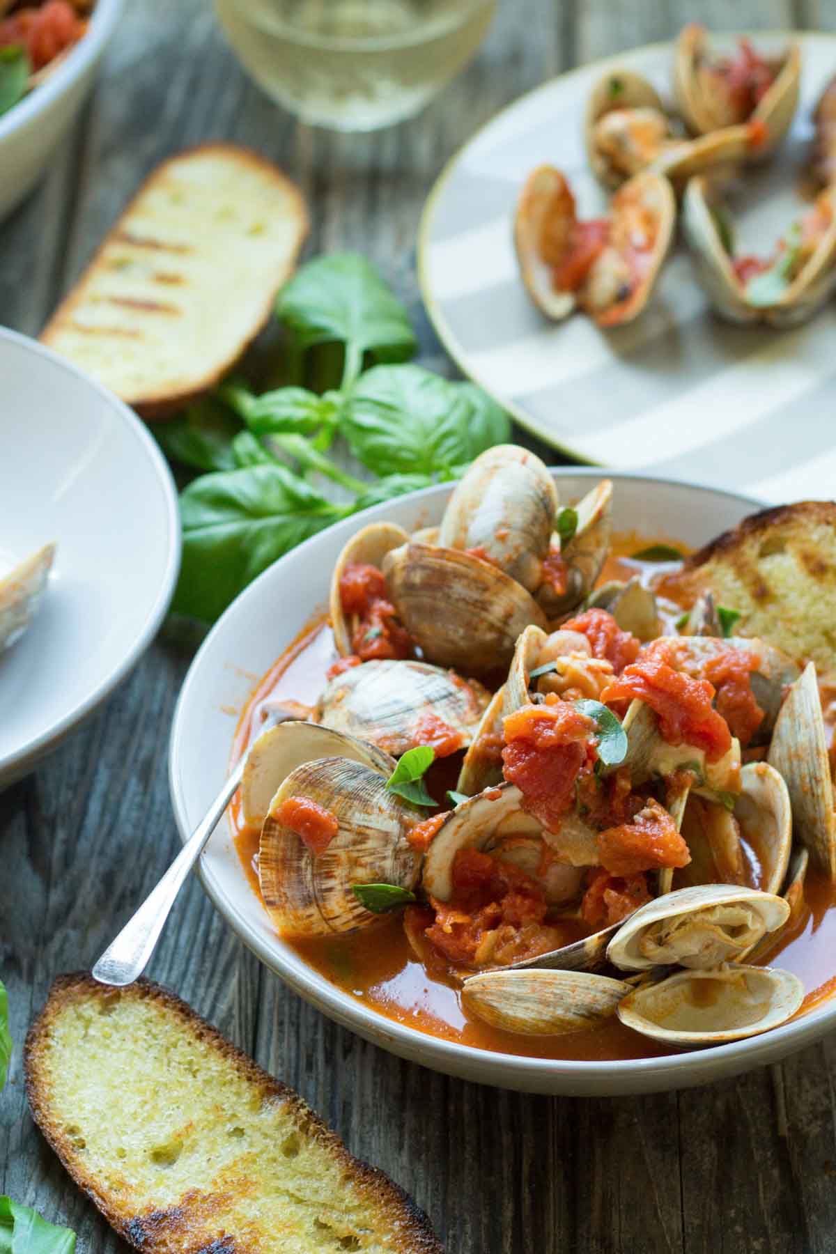 Wide shot of the dish surrounded by fresh basil leaves and glasses of white wine, showing clams in a chunky tomato sauce with herbs and toast.