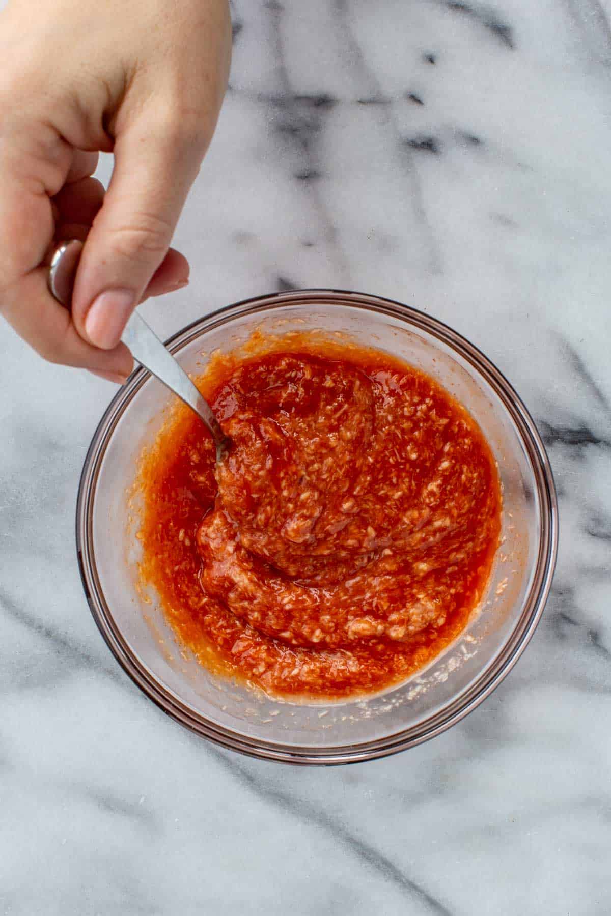 Hand stirring the cocktail sauce ingredients together in a glass bowl using a spoon.