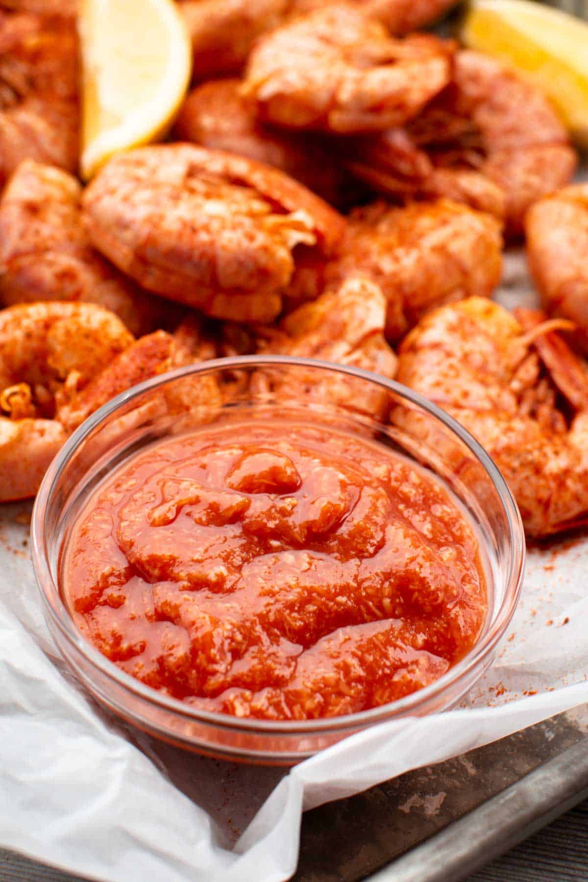 Glass bowl of cocktail sauce placed on a tray surrounded by seasoned shrimp and lemon wedges.