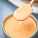 Spoon lifting smooth and creamy Creole remoulade sauce from a small ceramic bowl with a sandwich roll visible in the background.
