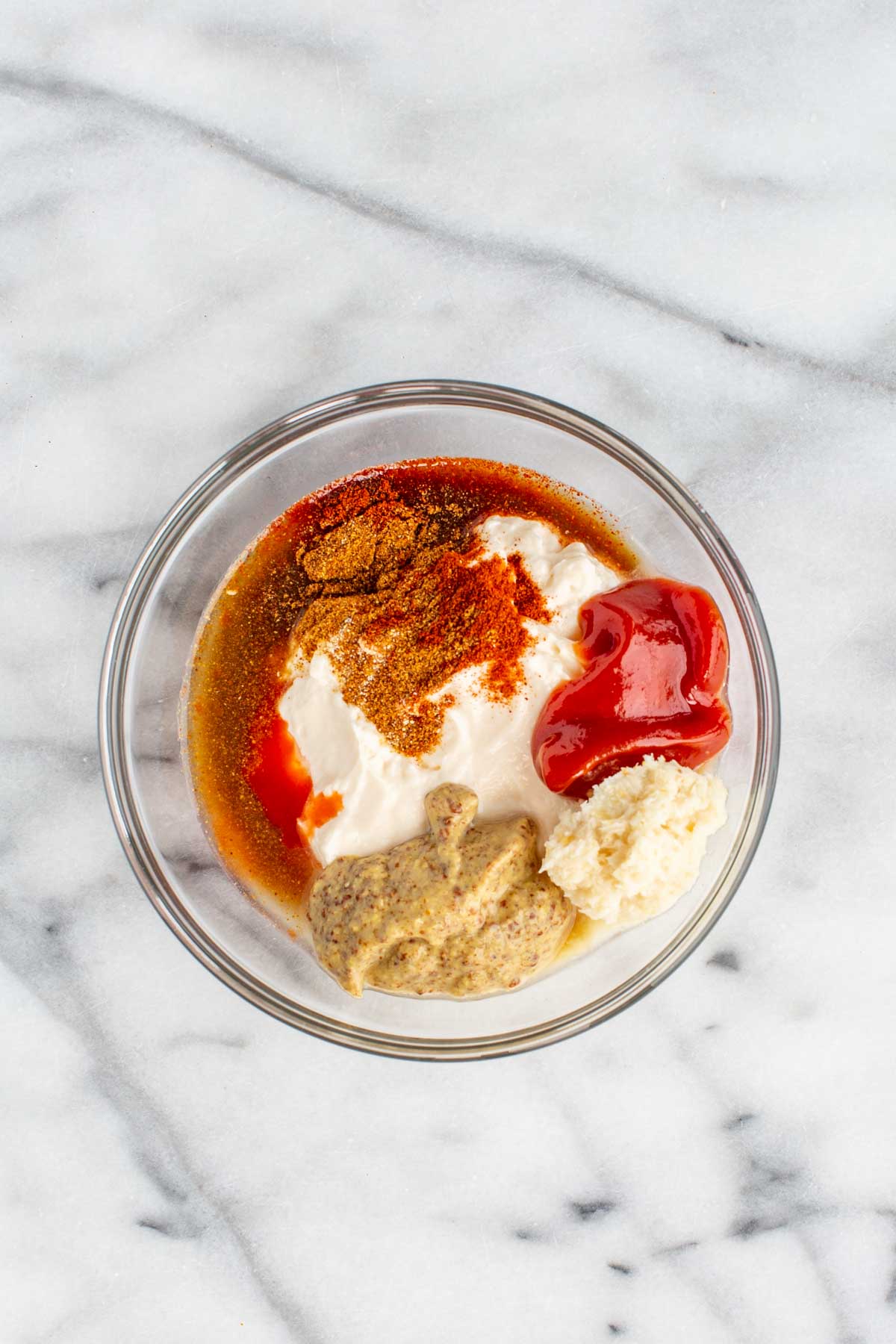 Overhead view of a glass bowl containing all the ingredients for Creole remoulade sauce before mixing, with swirls of mayonnaise, mustard, ketchup, spices, horseradish, and hot sauce visible.