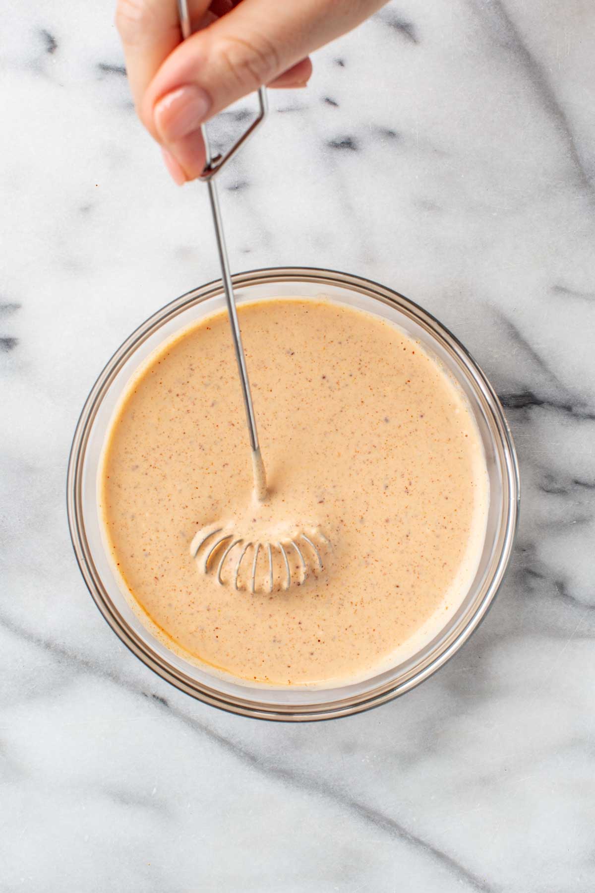 Close-up of a hand whisking together the Creole remoulade sauce ingredients in a glass bowl until smooth and creamy.