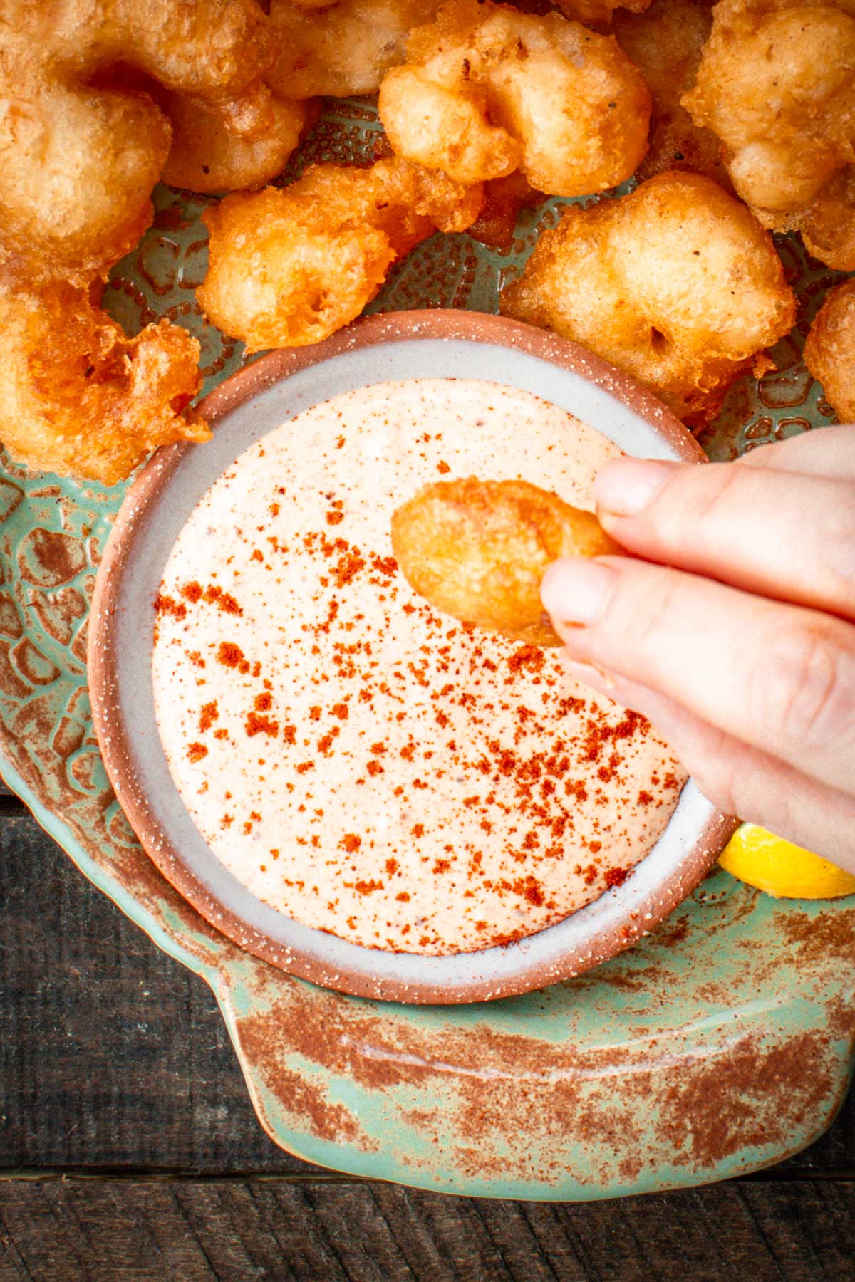 Hand dipping a fried shrimp into a bowl of paprika-dusted Creole remoulade sauce, surrounded by more shrimp on a rustic plate.