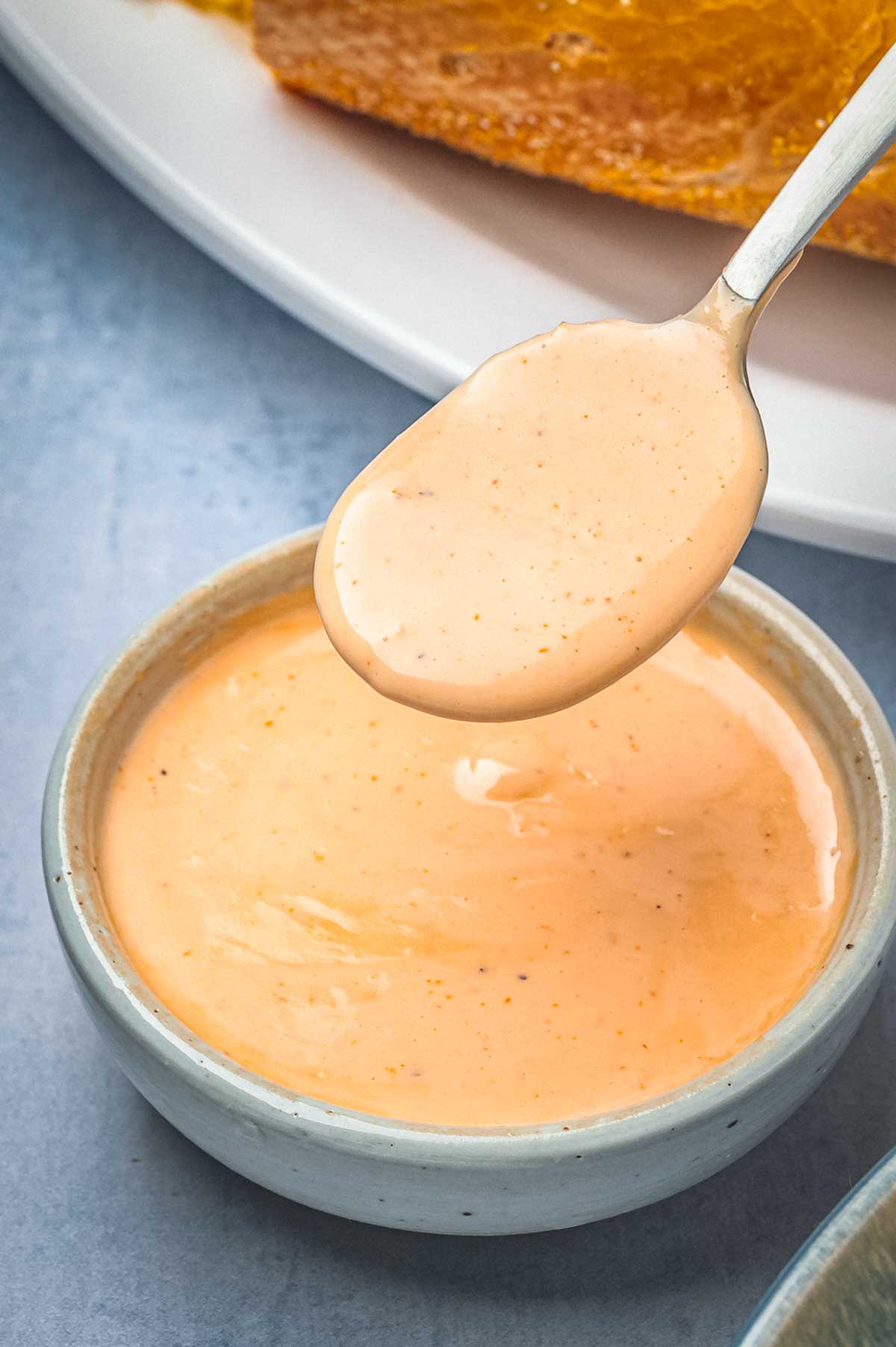 Spoon lifting smooth and creamy Creole remoulade sauce from a small ceramic bowl with a sandwich roll visible in the background.