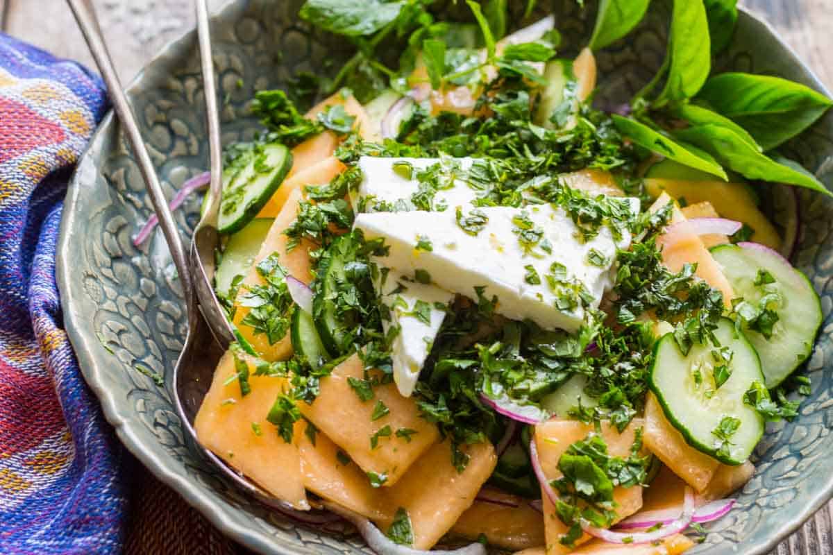Overhead view of a colorful cucumber melon salad in a patterned ceramic bowl, garnished with fresh basil and a large wedge of feta, surrounded by sliced cucumbers and red onions.