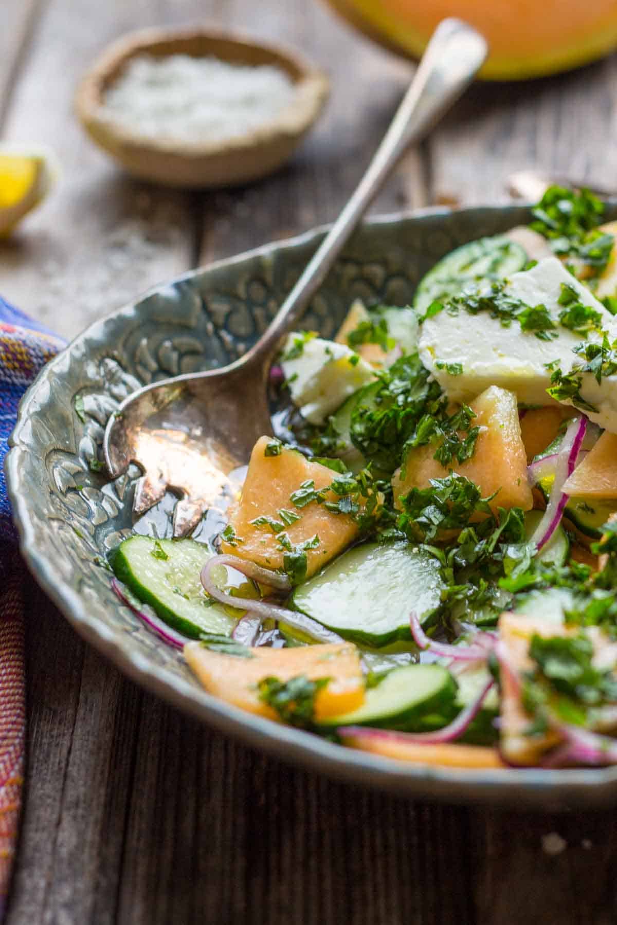 Side angle of cucumber melon salad in a green ceramic bowl with a serving spoon, highlighting the juicy cantaloupe, cucumbers, red onion, and fresh herb topping.
