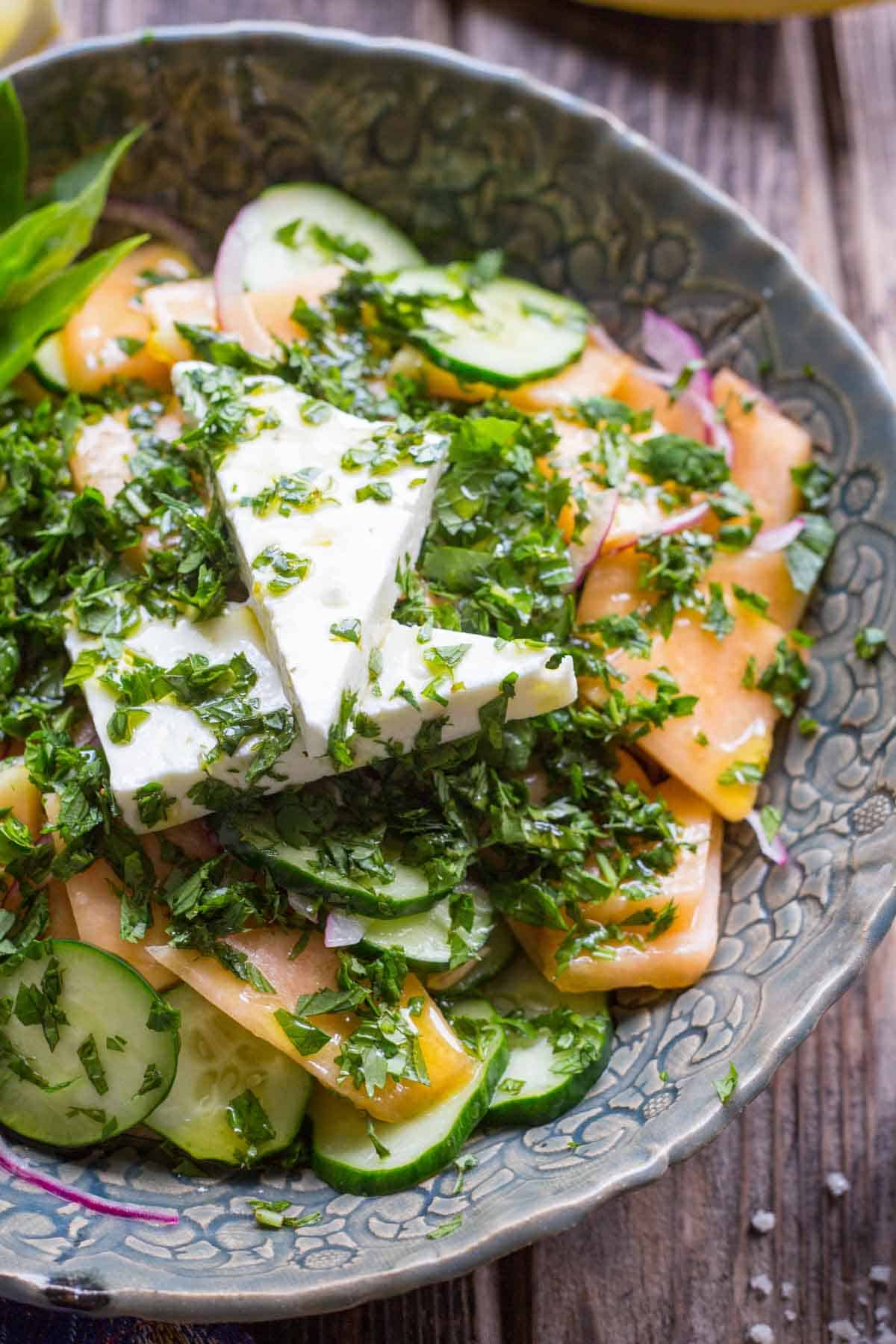 Top-down view of a vibrant salad made with sliced cantaloupe, cucumbers, and onions, topped with two stacked wedges of feta and a generous sprinkle of herbs in a decorative ceramic bowl.