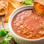 Close-up of salsa with chip dipped in: A white ramekin filled with tomato salsa, with a crispy tortilla chip dipped on the side.