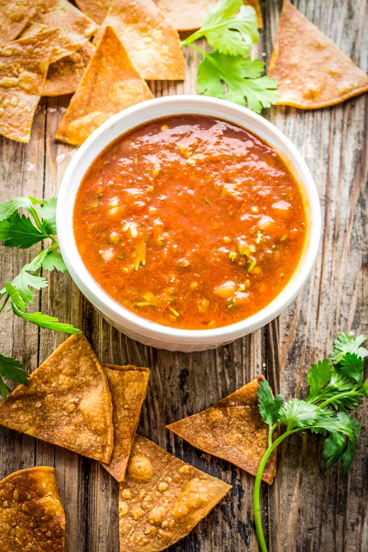 Overhead of salsa with chips and cilantro:
Bowl of tomato salsa on a rustic wood surface, surrounded by tortilla chips and fresh cilantro sprigs.