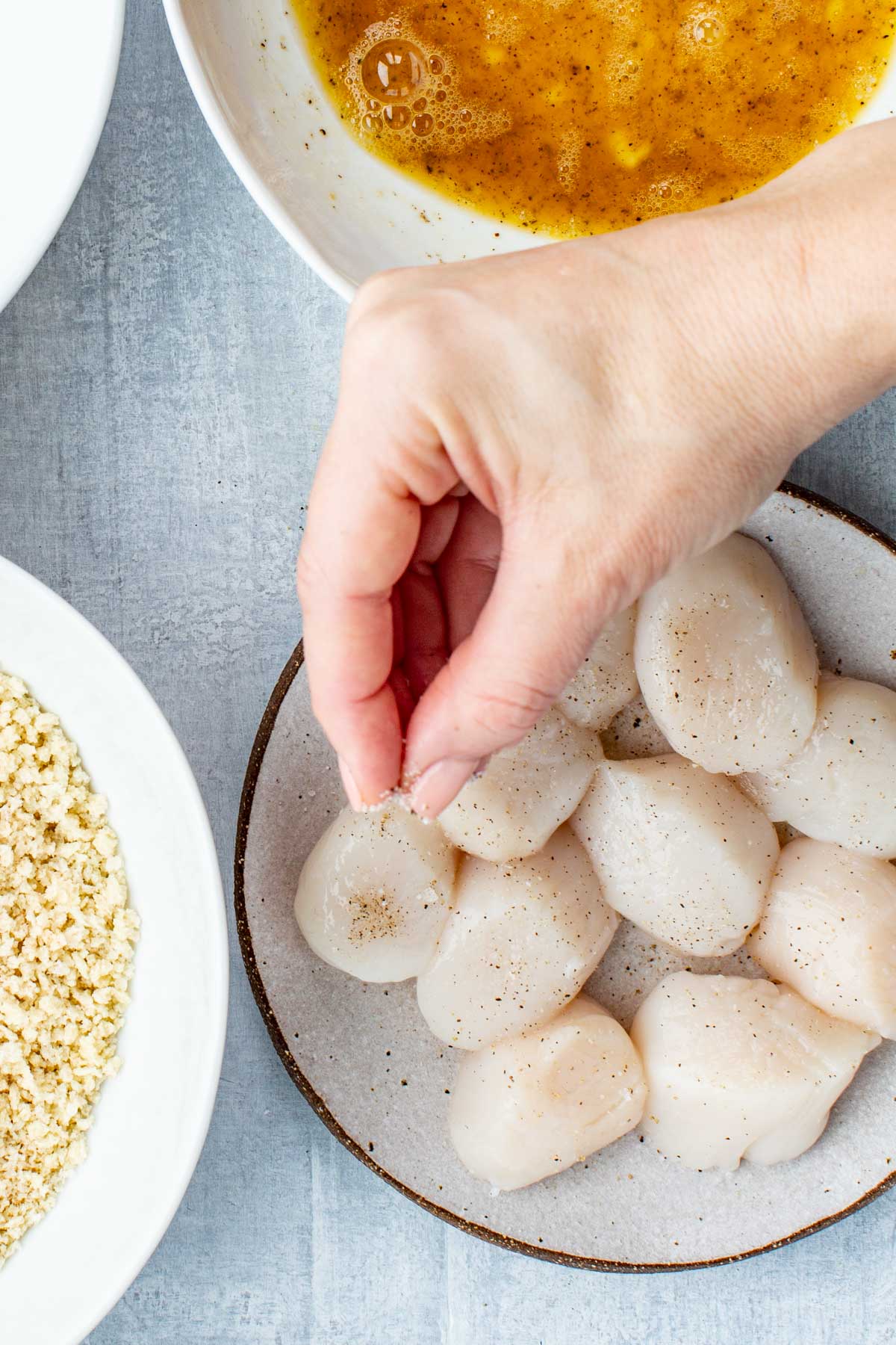 Hand sprinkling salt over a plate of scallops near bowls of egg wash, flour, and panko breadcrumbs.