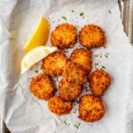 Fried scallops arranged on parchment paper with lemon wedges and chopped herbs.