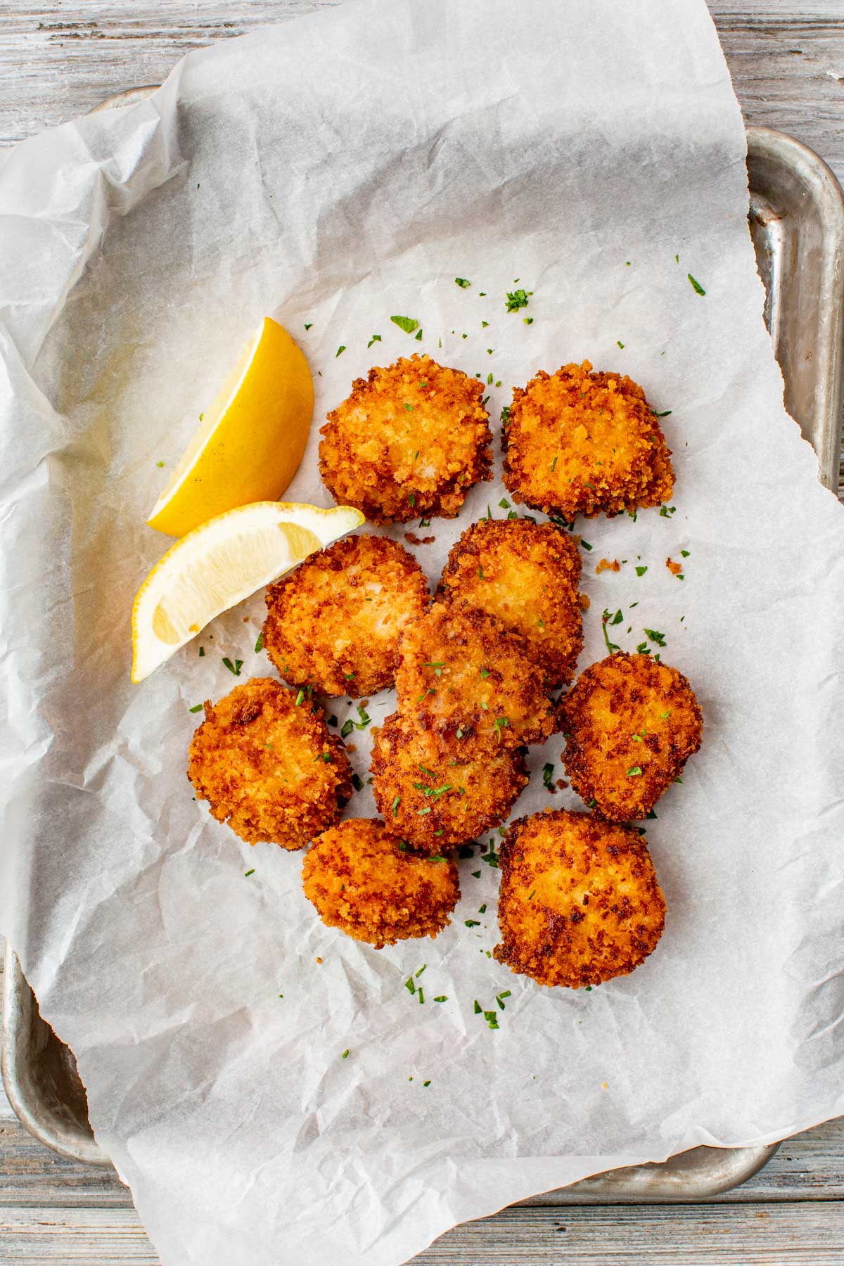 Fried scallops arranged on parchment paper with lemon wedges and chopped herbs.