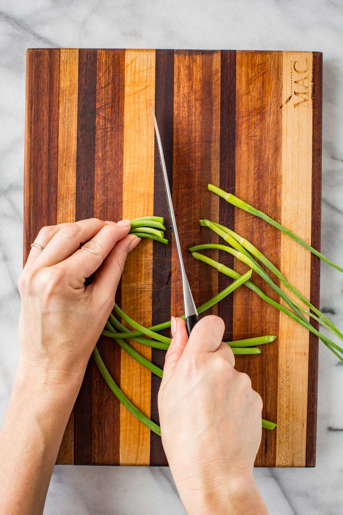 Close-up of hands using a sharp knife to cut fresh garlic scapes into small pieces on a two-tone wood cutting board.