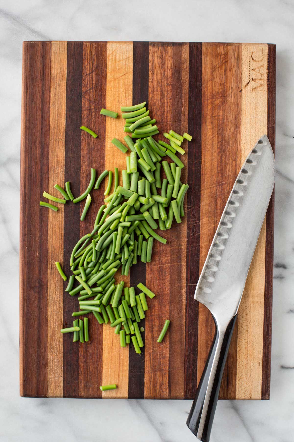 Top-down view of a pile of chopped garlic scapes next to a large chef’s knife on a striped cutting board.