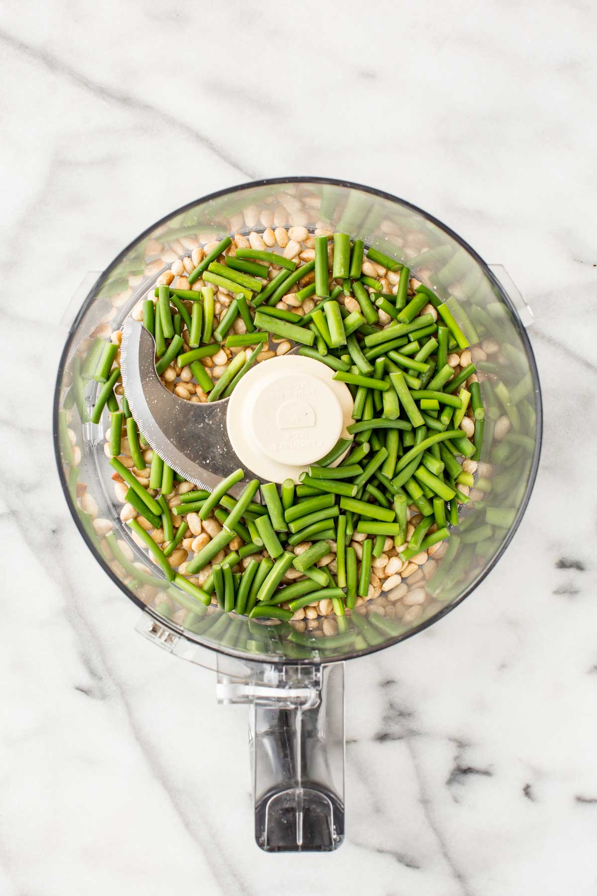 Top-down image of a food processor bowl filled with toasted pine nuts and vibrant green garlic scapes before blending.